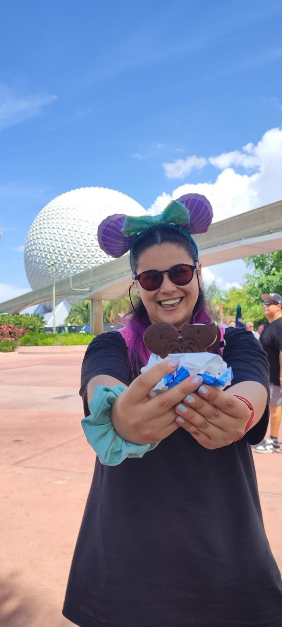 Coni sonriendo y sosteniendo un helado Mickey Ice Cream Sandwich frente a la icónica esfera Spaceship Earth en el parque EPCOT de Walt Disney World.